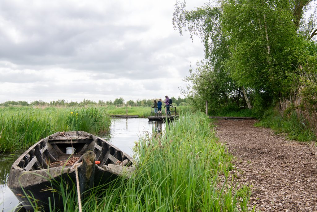 We zien een gezin op een trekpontje staan dat zich in een rietlandschap bevindt. Op de voorgrond zien we een open boot liggen.